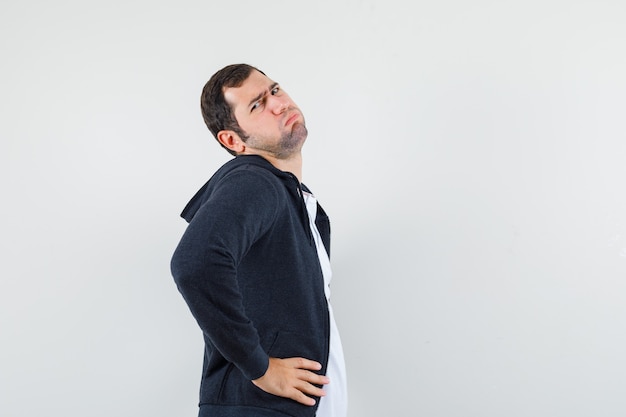 Junger Mann, der Hände auf Taille im T-Shirt, in der Jacke hält und gelangweilt schaut. .