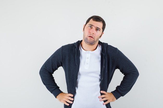 Junger Mann, der Hände auf Taille im T-Shirt, in der Jacke und in der verwirrten Vorderansicht hält.