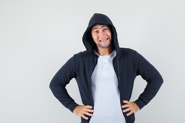 Junger Mann, der Hände auf Taille in Jacke, T-Shirt hält und fröhlich schaut. Vorderansicht.