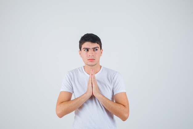 Junger Mann, der Hände in der Gebetsgeste im T-Shirt hält und hoffnungsvoll aussieht. Vorderansicht.