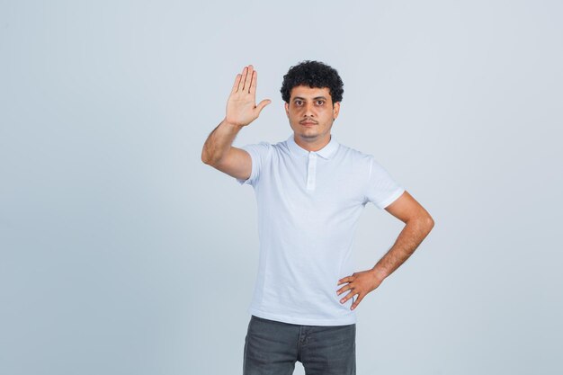 Junger Mann, der Hand an der Taille hält, während er Stoppschild in weißem T-Shirt und Jeans zeigt und ernst aussieht, Vorderansicht.