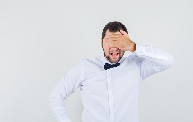 Junger Mann, der Hand auf Augen im weißen Hemd hält und aufgeregt, Vorderansicht schaut.