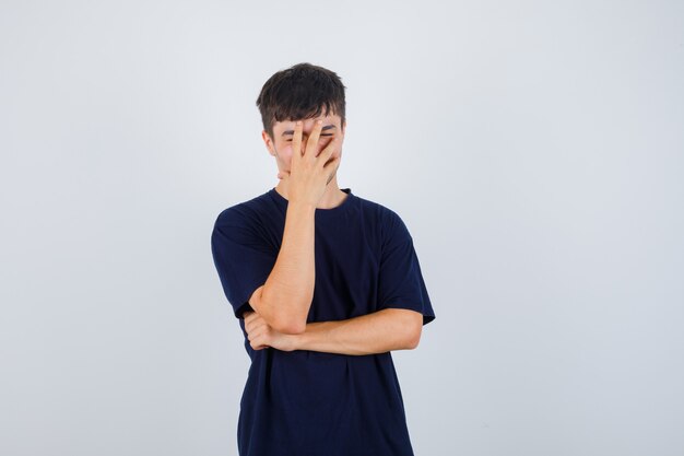 Junger Mann, der Hand auf Gesicht im schwarzen T-Shirt hält und aufgeregt schaut. Vorderansicht.