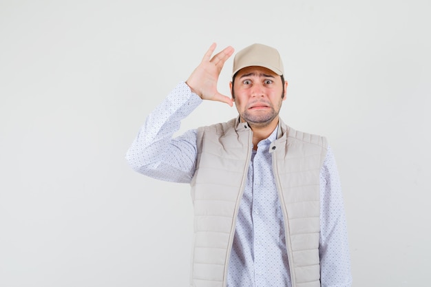 Junger Mann, der Hand nahe Ohr in beiger Jacke und Mütze erhebt und überrascht schaut. Vorderansicht.