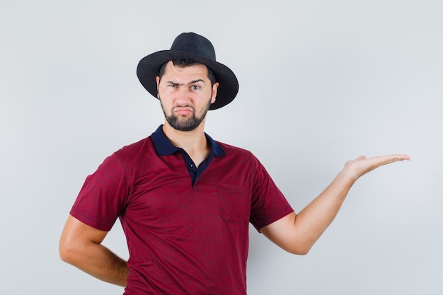 Junger Mann, der Handfläche beiseite spreizt, während er in T-Shirt, Hut, Vorderansicht finster blickt.