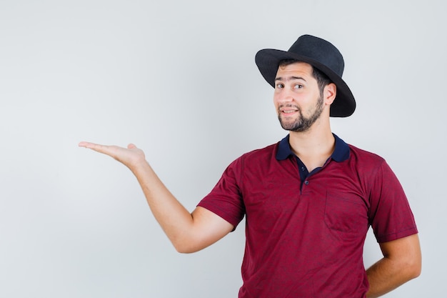 Junger Mann, der Handfläche in T-Shirt, Hut und fröhlich aussehend ausbreitet. Vorderansicht.