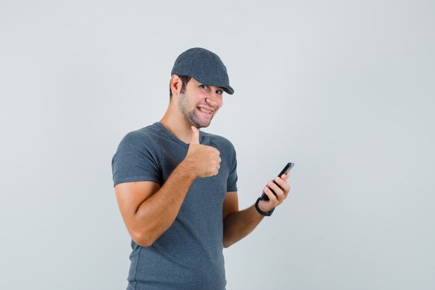 Junger Mann, der Handy hält, zeigt Daumen oben in der T-Shirt-Kappe und schaut lustig