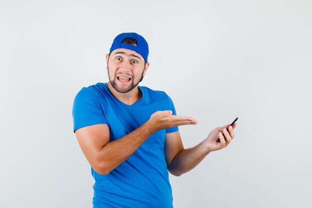 Junger Mann, der Handy im blauen T-Shirt und in der Kappe zeigt und verwirrt schaut