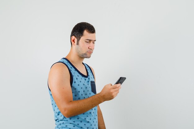 Junger Mann, der Handy im blauen Unterhemd benutzt und beschäftigt schaut. Vorderansicht.