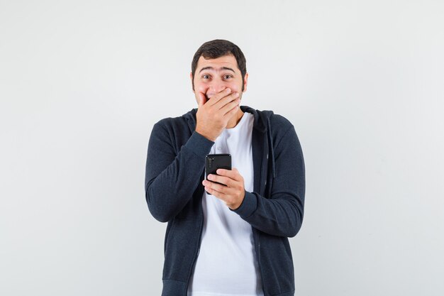 Junger Mann, der Handy im T-Shirt, in der Jacke hält und froh schaut. Vorderansicht.