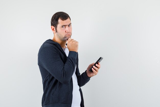 Junger Mann, der Handy im T-Shirt, in der Jacke hält und nachdenklich, Vorderansicht schaut.