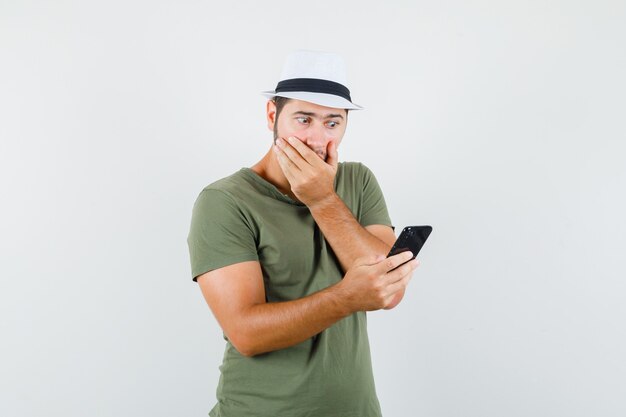 Junger Mann, der Handy in grünem T-Shirt und Hut betrachtet und überrascht schaut