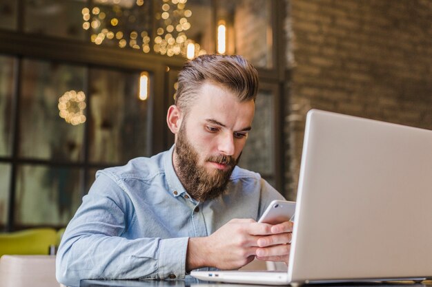 Junger Mann, der Handy mit Laptop auf Schreibtisch verwendet