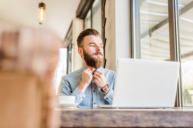 Junger Mann, der Hemd mit Tasse Kaffee-Anzeigenlaptop auf Schreibtisch knöpft