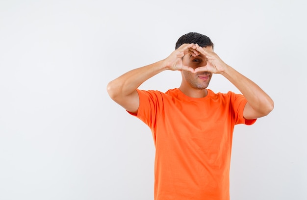 Junger Mann, der Herzgeste im orange T-Shirt zeigt und lustig schaut