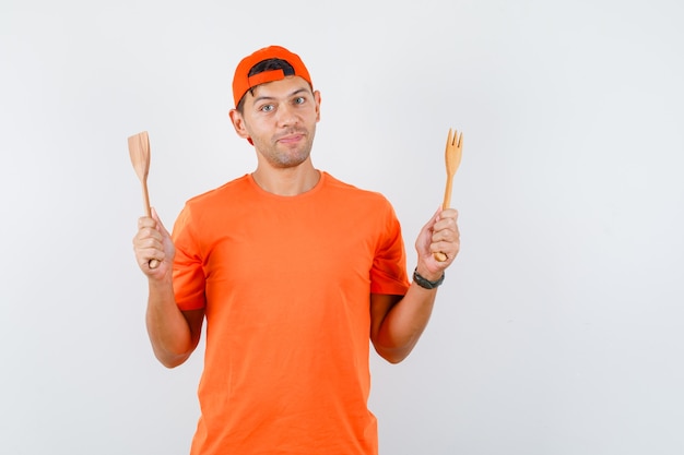 Junger Mann, der hölzerne Gabel und Spatel im orange T-Shirt und in der Kappe hält und fröhlich schaut