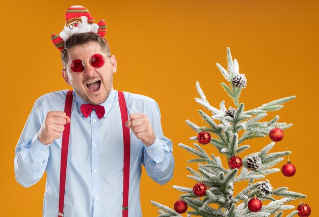 Junger Mann, der Hosenträgerfliege im Rand mit Weihnachtsmann und roter Brille trägt, die neben Weihnachtsbaum glücklich und aufgeregt über orange Wand stehen