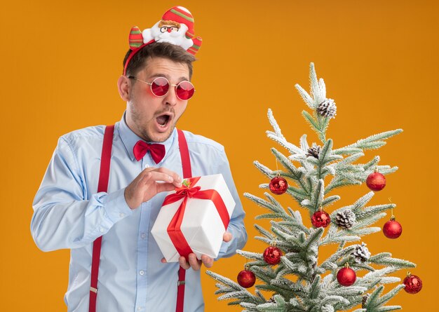 Junger Mann, der Hosenträgerfliege im Rand mit Weihnachtsmann und roter Brille trägt, steht neben Weihnachtsbaum, der ein Geschenk glücklich und aufgeregt über orange Hintergrund hält