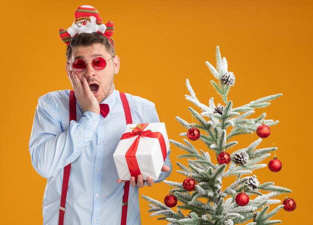 Junger Mann, der Hosenträgerfliege im Rand mit Weihnachtsmann und roter Brille trägt, steht neben Weihnachtsbaum, der ein Geschenk glücklich und aufgeregt über orange Hintergrund hält