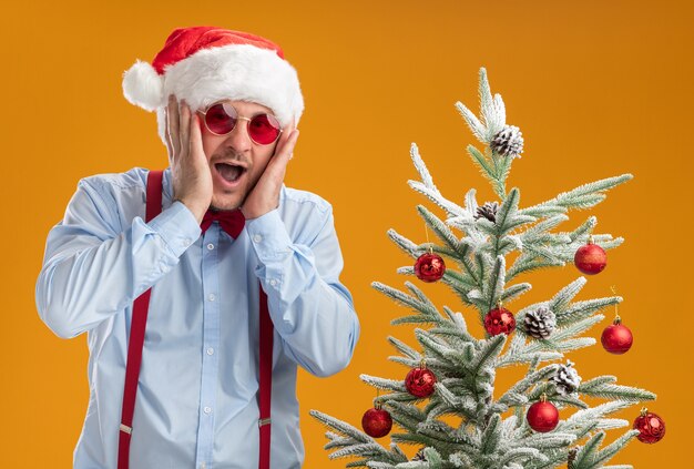 Junger Mann, der Hosenträgerfliege im Weihnachtsmannhut und in der roten Brille trägt, erstaunt und überrascht, nahe Weihnachtsbaum über orange Wand stehend