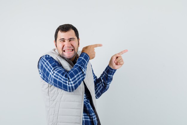 Junger Mann, der im Hemd, in der ärmellosen Jacke beiseite zeigt und lustig schaut. Vorderansicht.