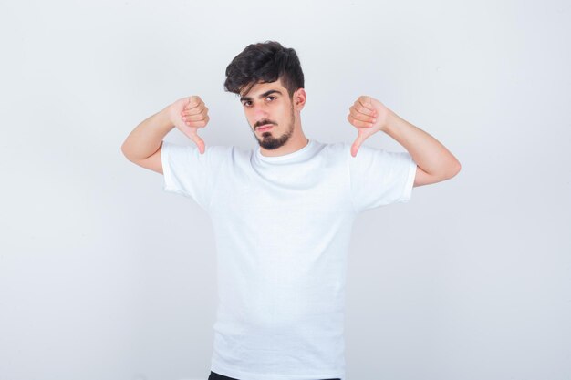 Junger Mann, der im weißen T-Shirt doppelte Daumen nach unten zeigt und selbstbewusst aussieht