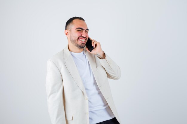 Junger Mann, der im weißen T-Shirt, in der Jacke mit dem Telefon spricht und glücklich aussieht. Vorderansicht.