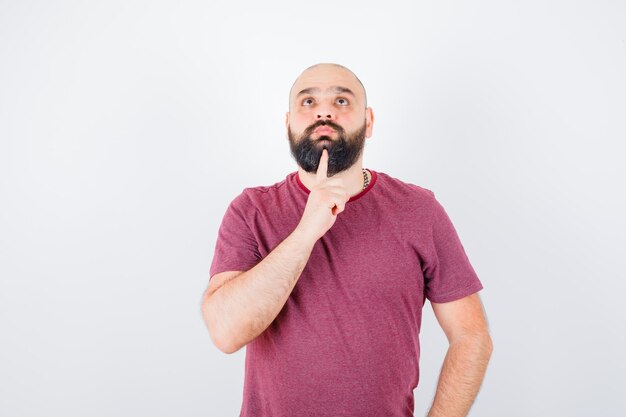 Junger Mann, der in denkender Pose im rosa T-Shirt steht und nachdenklich aussieht. Vorderansicht.