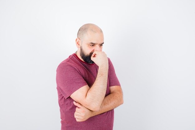 Junger Mann, der in denkender Pose im rosa T-Shirt steht und nachdenklich aussieht. Vorderansicht.