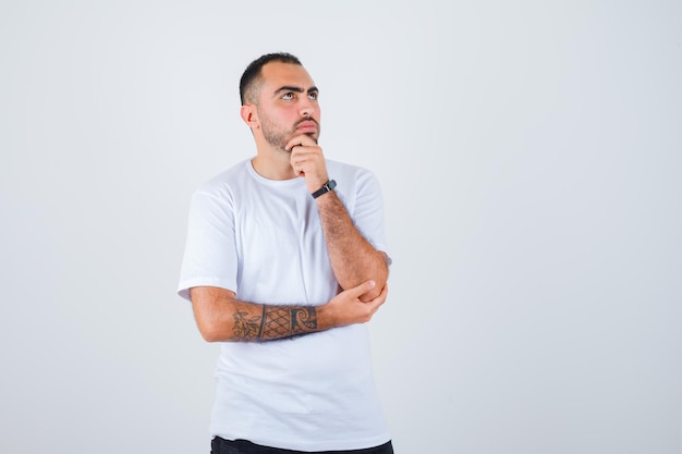 Junger Mann, der in denkender Pose in weißem T-Shirt und schwarzer Hose steht und nachdenklich aussieht