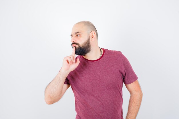 Junger Mann, der in denkender Pose steht, im rosa T-Shirt wegschaut und nachdenklich aussieht. Vorderansicht.