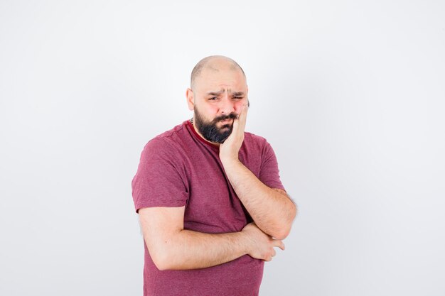 Junger Mann, der in denkender Pose steht, sich in rosa T-Shirt auf die Handfläche lehnt und nachdenklich aussieht. Vorderansicht.