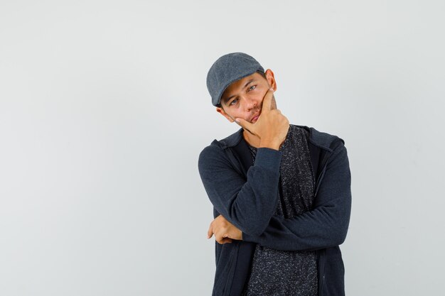 Junger Mann, der in der denkenden Pose im T-Shirt, in der Jacke, in der Mütze steht und ruhig schaut. Vorderansicht.