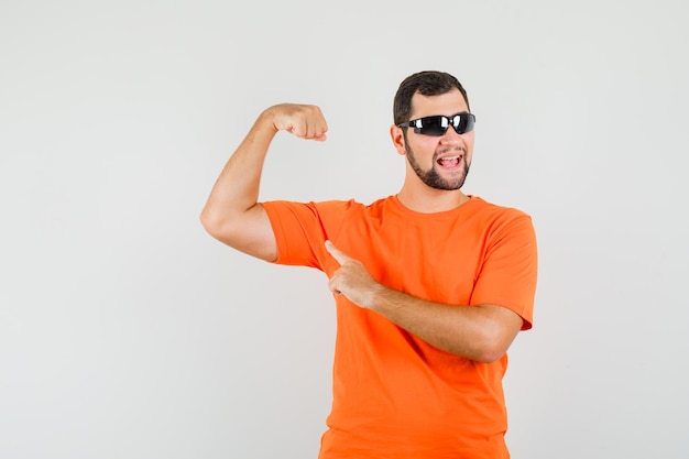 Junger Mann, der in orangefarbenem T-Shirt auf Armmuskeln zeigt und selbstbewusst aussieht. Vorderansicht.