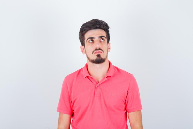 Junger Mann, der in rosa T-Shirt nach oben schaut und nachdenklich aussieht, Vorderansicht.