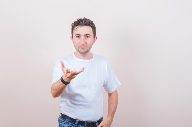 Junger Mann, der in weißem T-Shirt, Jeans und verwirrtem Blick eine Frage-Geste macht