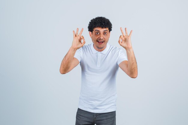 Junger Mann, der in weißem T-Shirt und Jeans gute Zeichen zeigt und glücklich aussieht, Vorderansicht.