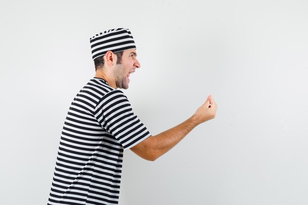 Junger Mann, der italienische Geste in T-Shirt, Hut tut und wütend schaut.
