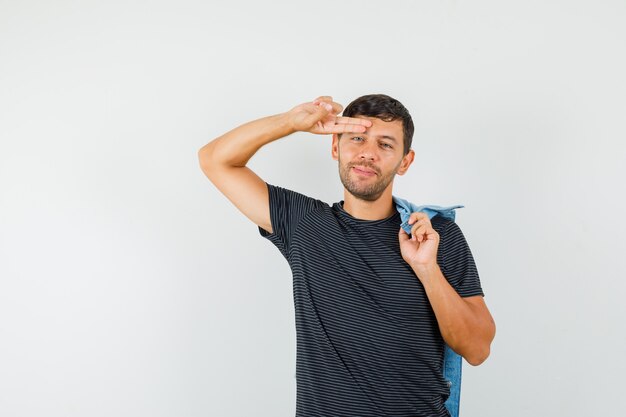 Junger Mann, der Jacke auf Rücken hält, Finger auf Stirn in T-Shirt hält und lustig schaut