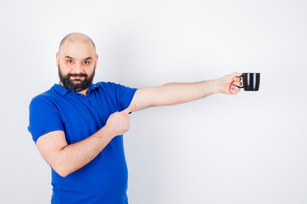 Junger mann, der jemandem eine tasse gibt, während er im blauen hemd darauf zeigt und sicher aussieht. .