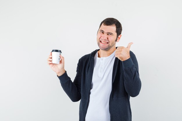 Junger Mann, der Kaffeetasse zum Mitnehmen hält und Daumen in weißem T-Shirt und schwarzem Kapuzenpullover mit Reißverschluss vorne zeigt und glücklich aussieht. Vorderansicht.