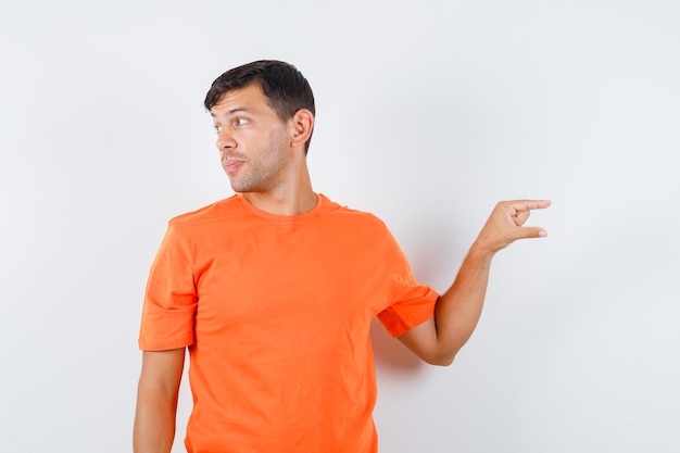 Junger Mann, der kleines Zeichen zeigt, während er im orangefarbenen T-Shirt beiseite schaut und konzentriert schaut