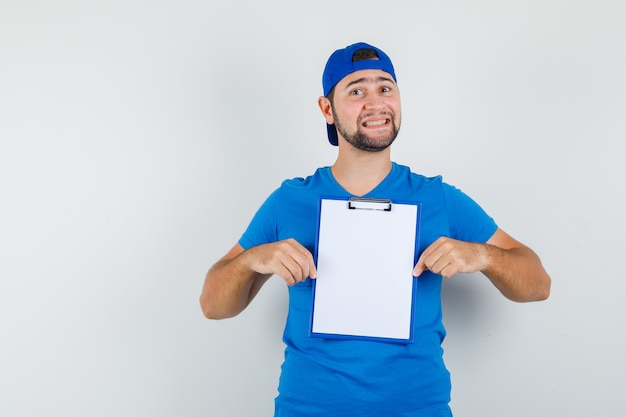 Junger Mann, der Klemmbrett im blauen T-Shirt und in der Kappe hält und fröhlich schaut