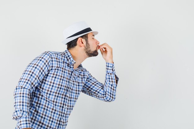 Junger Mann, der leckere Geste zeigt, indem er Finger in kariertem Hemd, Hut küsst und entzückt aussieht. Vorderansicht.