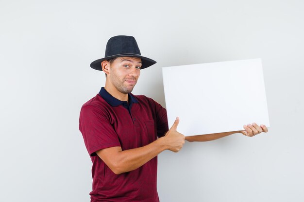 Junger Mann, der leere Leinwand mit Daumen hoch im T-Shirt, im Hut hält und fröhlich schaut. Vorderansicht.