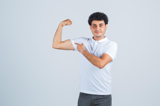 Junger Mann, der Machtgeste in weißem T-Shirt und Jeans zeigt und mächtig aussieht, Vorderansicht.