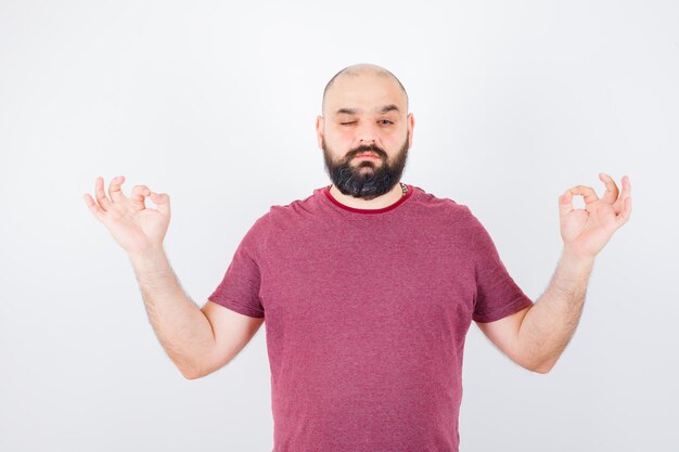 Junger Mann, der Meditation tut, während er in rosa T-Shirt zwinkert und ruhig aussieht. Vorderansicht.