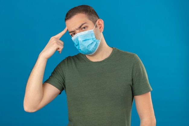 Junger Mann, der medizinische Gesichtsmaske trägt, die seinen Kopf mit dem auf Blau lokalisierten Finger berührt