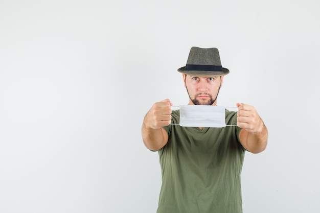 Kostenloses Foto junger mann, der medizinische maske im grünen t-shirt, im hut hält und ernst schaut