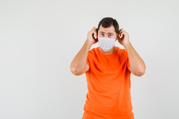 Junger Mann, der medizinische Maske im orange T-Shirt trägt und vorsichtig schaut, Vorderansicht.